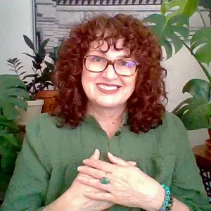 A photographic image of me from a couple of years ago. I am surrounded by houseplants, wearing a pretty green top, and have my hands crossed on my heart center. I am smiling broadly and my energy feels authentic, warm, and open. My material life has changed. I no longer live in that apartment or have those plants, but I am still authentic, warm, and open to all that is meant for me.