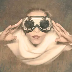 A vintage looking photograph of a woman. The perspective is from overhead looking down at her. She is looking up into a bright light and wearing big round goggles with big black lenses. Her hands are near here goggles, and she is focusing on what she is seeing. It's almost as if she is seeing into the future.