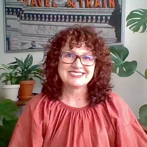 A photograph of me. I have a big, warm smile on my face and am wearing a terracotta-colored top. My auburn, curly hair is cut into a shag. In the background are plants and some artwork.