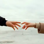 Two hands reaching towards each other. We see they are wearing jackets and autumn sweaters. The background is a grey, cold, seascape.