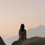 A figure sitting on large rocks and looking toward mountains in muted shades of lavander.. The sky is muted shades of pink as if it is either dawn or dusk.