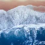 An overhead image of blue sea and white foam waves crashing onto a sandy beach.