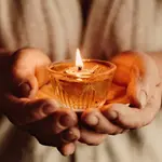 A pair of hand cupping a glass dish with a lit candle casting soft light. In the background are the gentle folds of a robe made of natural fibers.