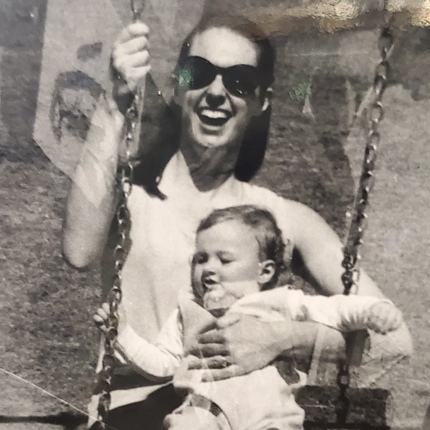 A very old photo of my mom on a playground swing with infant me in her arms.