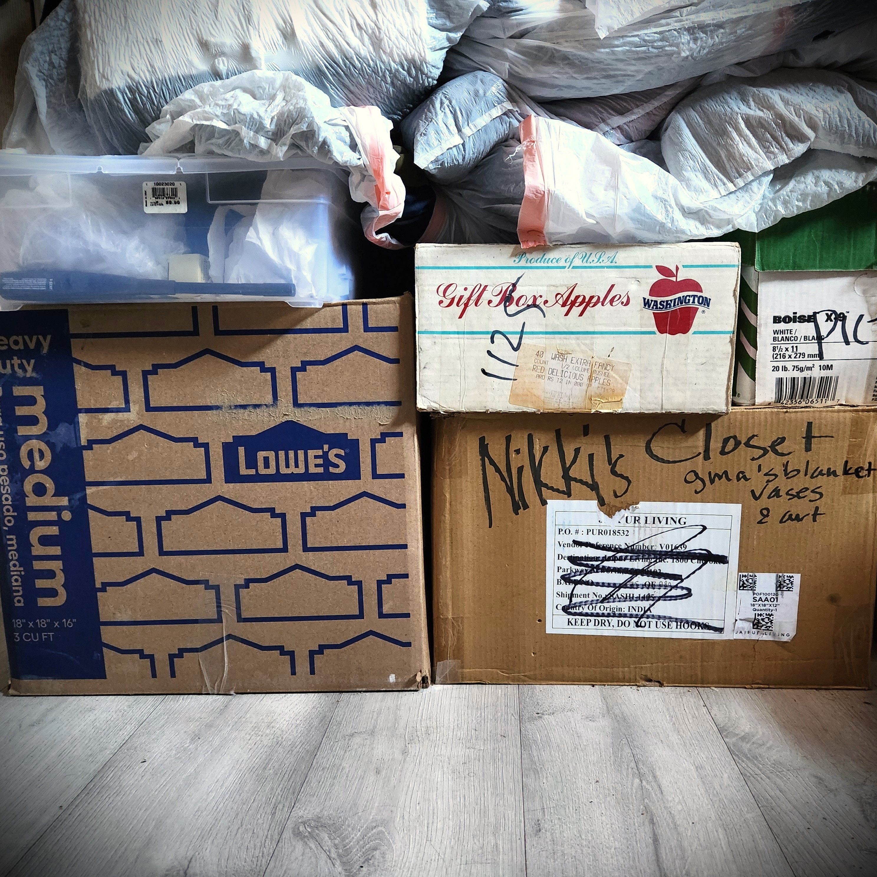 A photograph of the boxes mentioned in the blog post. From left to right: a Lowe's box with "medium" on the left side; to the right of it a box with red script that says, "Gift Box Apples"; below that one a box with handwriting that says, "Nikki's Closet". The boxes sit on gray vinyl flooring. They are topped with one smaller plastic box and many plastic bags. A photograph of the boxes mentioned in the blog post. From left to right: a Lowe's box with "medium" on the left side; to the right of it a box with red script that says, "Gift Box Apples"; below that one a box with handwriting that says, "Nikki's Closet". The boxes sit on gray vinyl flooring. They are topped with one smaller plastic box and many plastic bags.