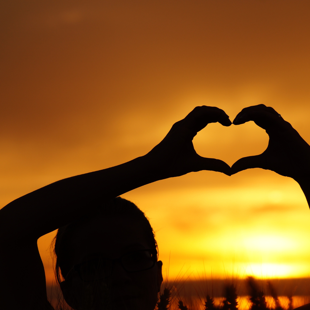 A person holding their hands above their head. Their fingers are making a heart shape and are silhouetted against a deeply colored sunrise.
