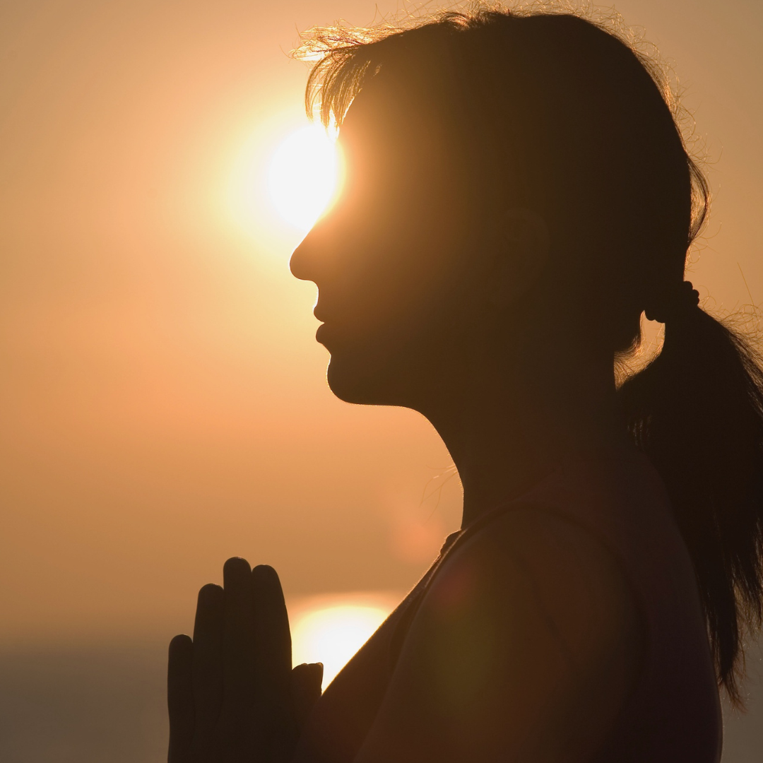 A photographic image of a person with their hands pressed together at their heart. They are silhouetted against a low sun. It could be either sunrise or sunset. We get to choose how we interpret the image.