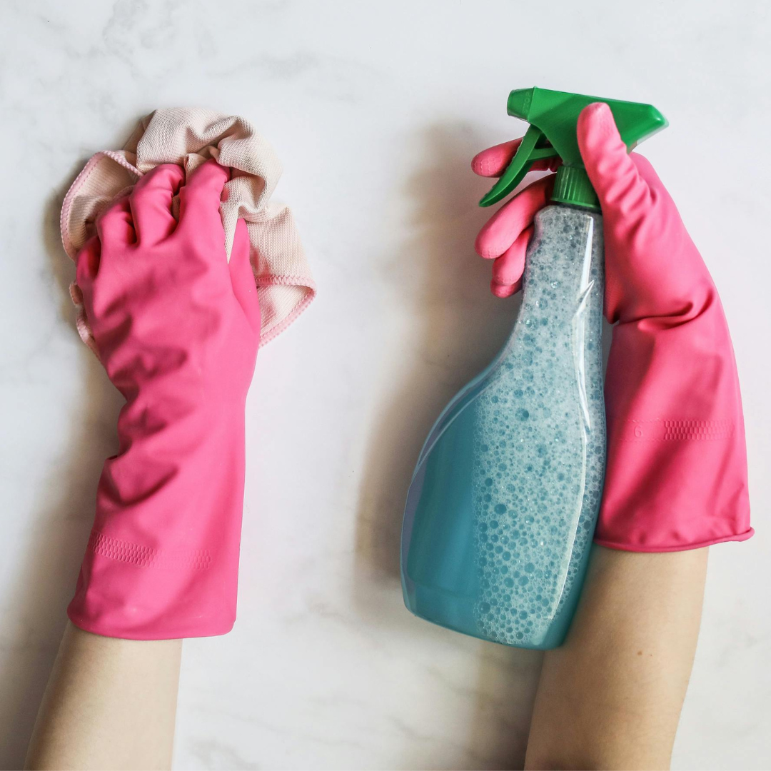 A photographic image of two hands in dark pink gloves. In the left hand is a white wash rag, and in the right hand a blue spray bottle of bubbly cleaner. The background is white lightly veined marble. A photographic image of two hands in dark pink gloves. In the left hand is a white wash rag, and in the right hand a blue spray bottle of bubbly cleaner. The background is white lightly veined marble.
