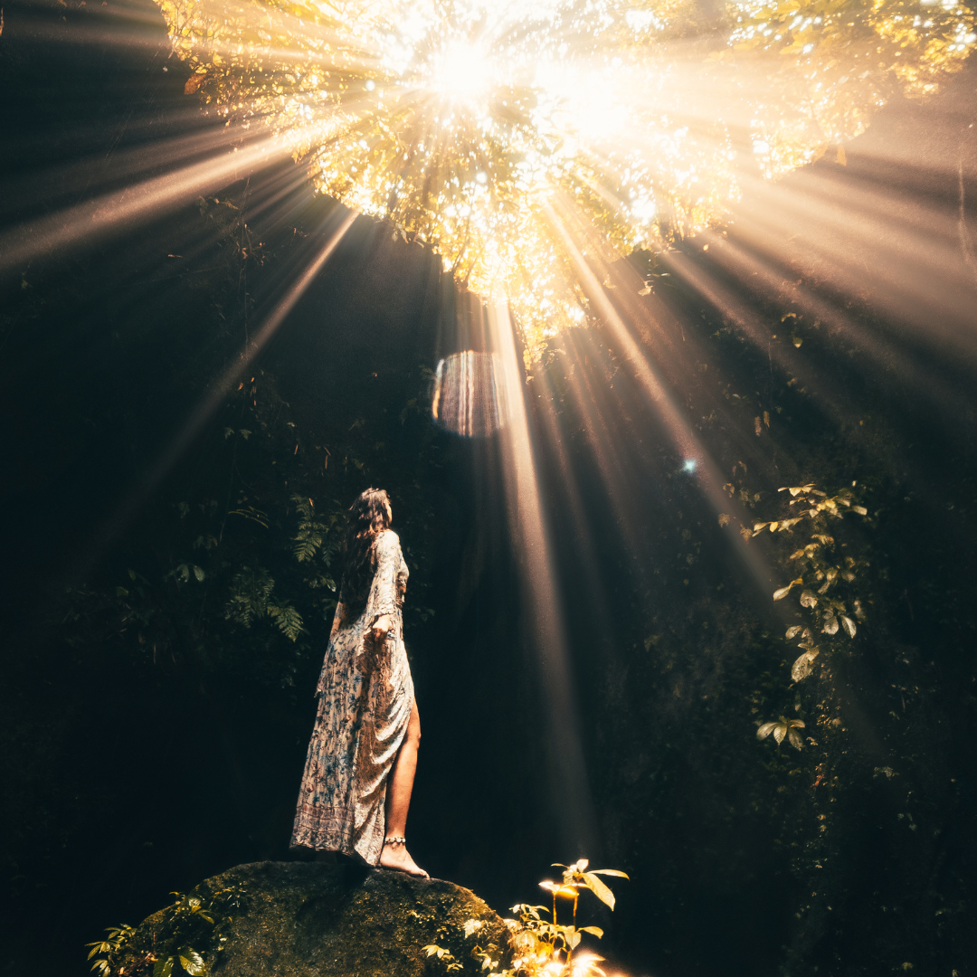 The image is an ethereal moment. A female figure in a flowing gown is looking up out of a dark cave and into the light of the sun. The trees surrounding the cave are creating a dappled effect. The sun is streaming in with brilliant rays of sunshine.