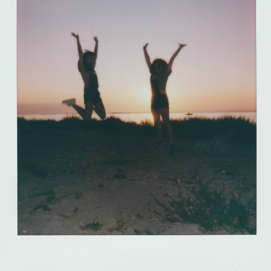 A vintage polaroid picture of two people, on the beach, jumping up with their arms held high as if in celebration. They are silhouetted on a background of a sunset on the ocean.
