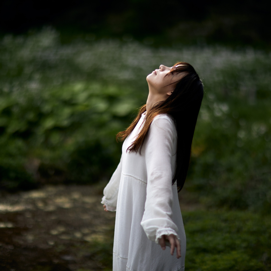 A woman standing in a forest. Wearing a white dress. Her arms are open and her face is relaxed and open to the sky.