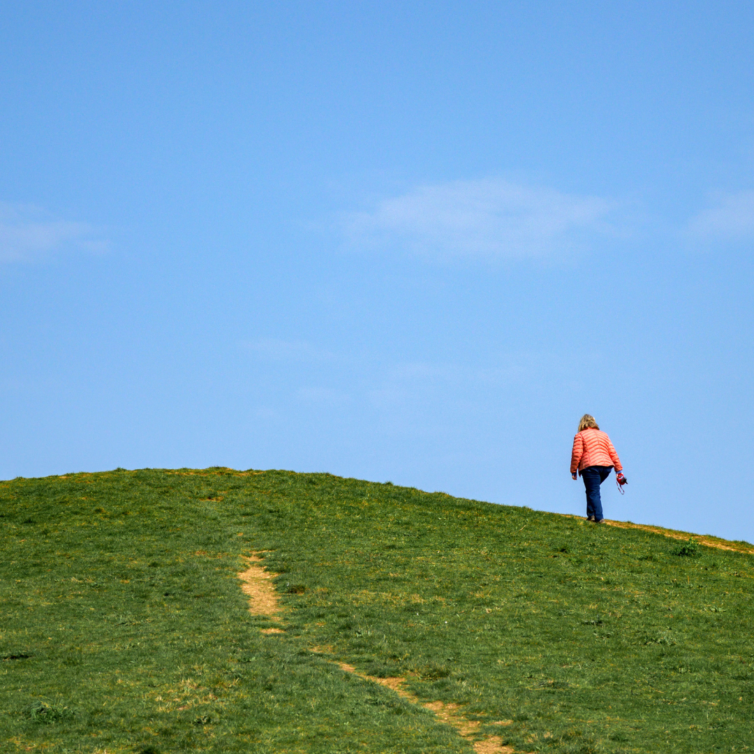 The image is of a woman walking up a hill. There is green grass and a clear blue sky. The image is of a woman walking up a hill. There is green grass and a clear blue sky.