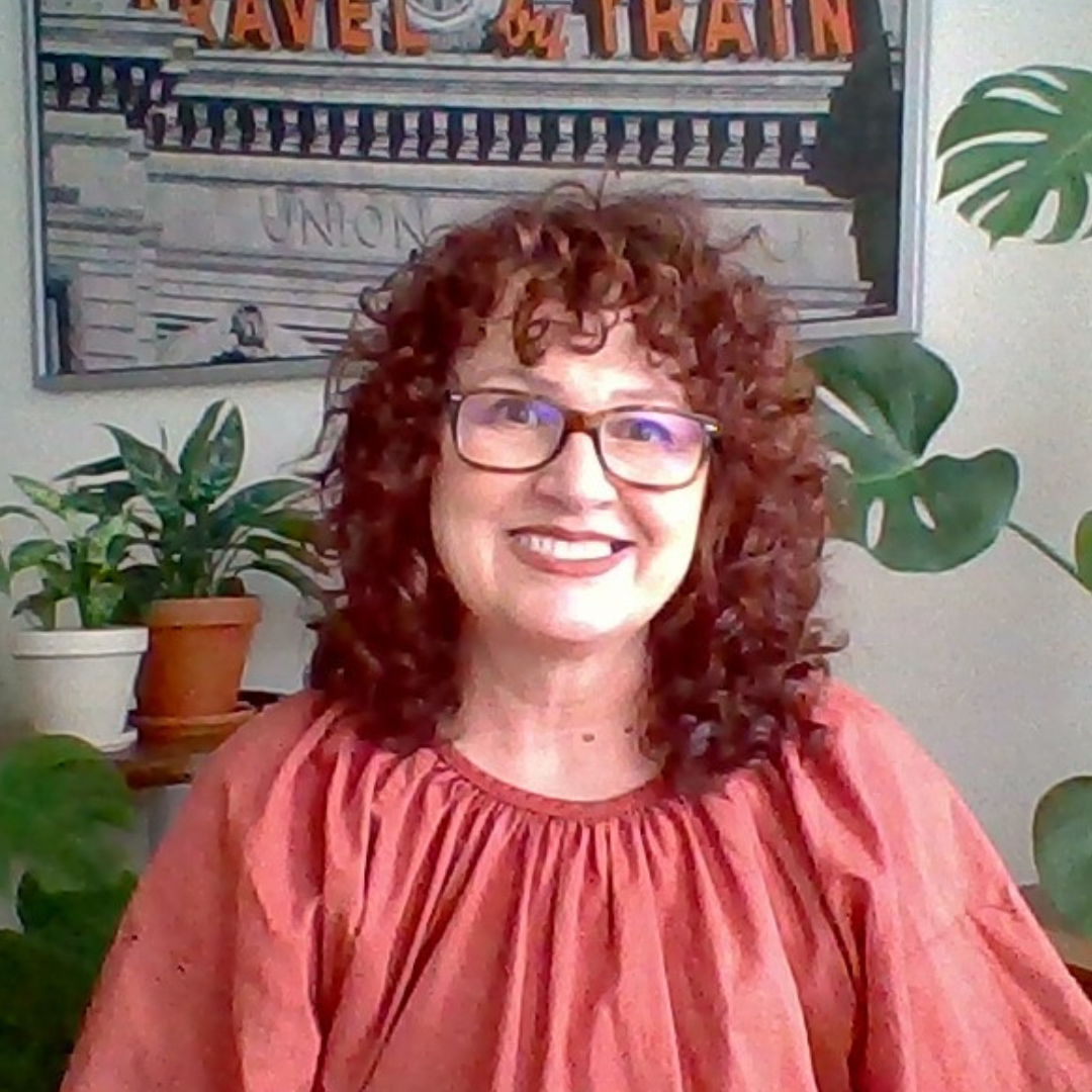 A photograph of me. I have a big, warm smile on my face and am wearing a terracotta-colored top. My auburn, curly hair is cut into a shag. In the background are plants and some artwork.