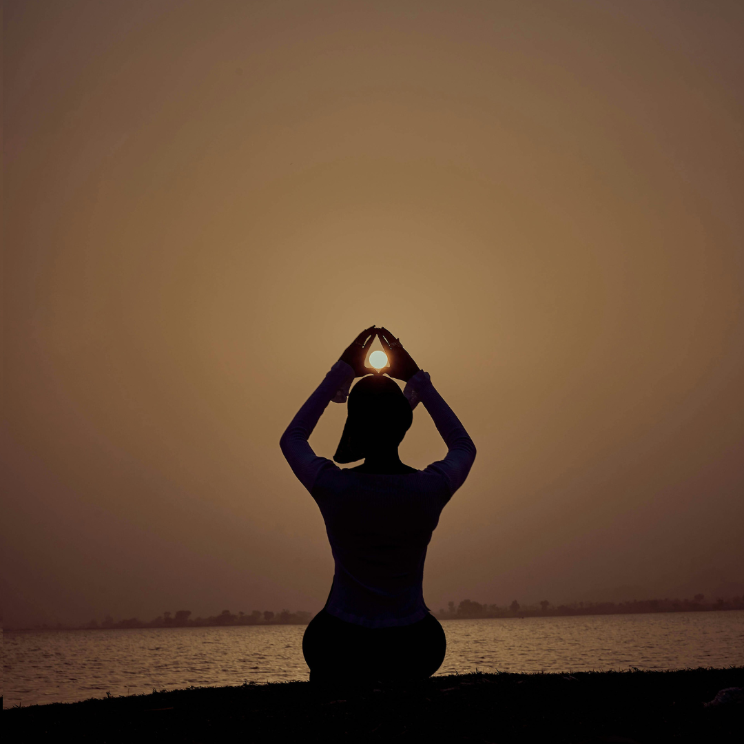 A woman silhouetted against the light of a low hanging moon. Her arms are raised above her head, and she appears to be holding the moon in her hands. The color palette is of warm browns.