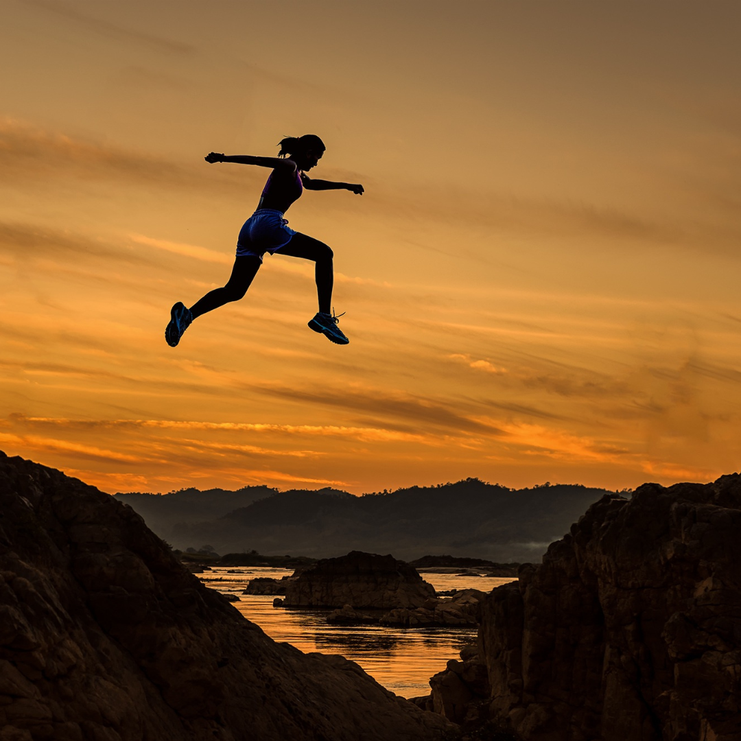 A woman making a huge leap on a background of a gold and orange sunset.