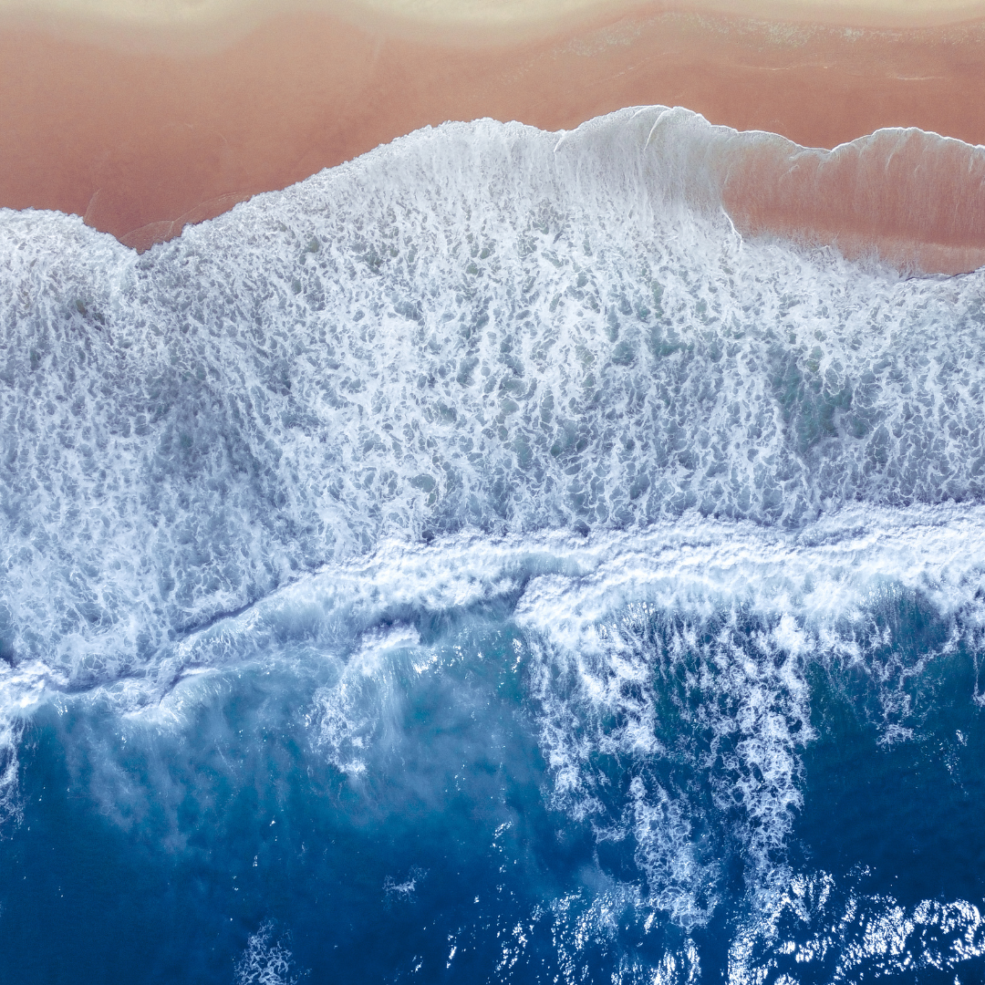 An overhead image of blue sea and white foam waves crashing onto a sandy beach. An overhead image of blue sea and white foam waves crashing onto a sandy beach.