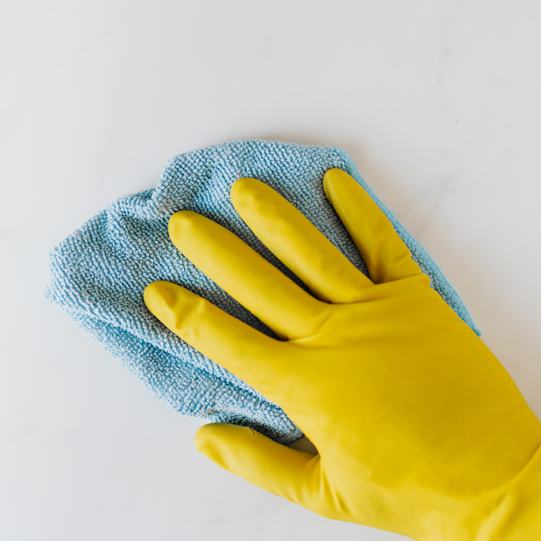 A photographic image of a hand wearing a yellow cleaning glove, holding a sky-blue cleaning cloth, swiping across a clean, white countertop.