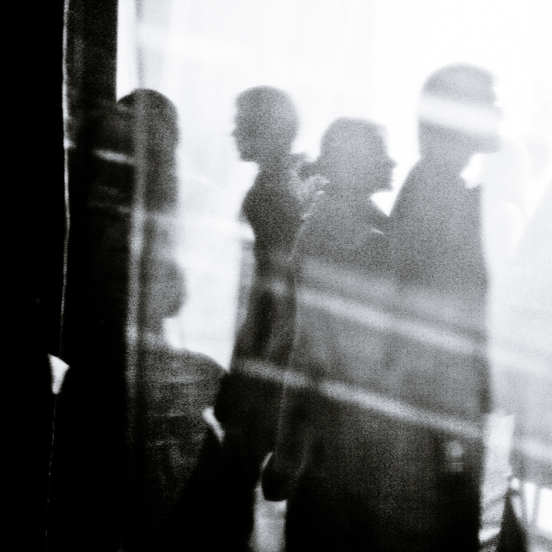 A somewhat abstract black and white image of people walking past a window. It is a study of dark and light.